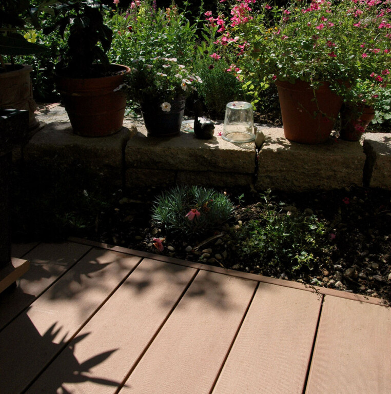 MYDECK Barfussdielen aus Holz Kunststoff (Polyethylen). Mediterrane Bepflanzung, Natursteinmäuerchen, hellbrauner WPC Balkonbelag bei Sonnenschein.