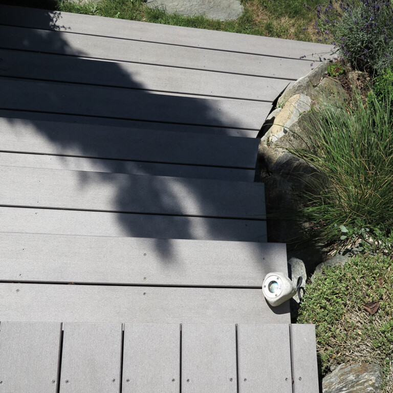 Graue WPC Terrasse mit WPC Stufen rund um Mauer geschnitten die an den Garten grenzt.