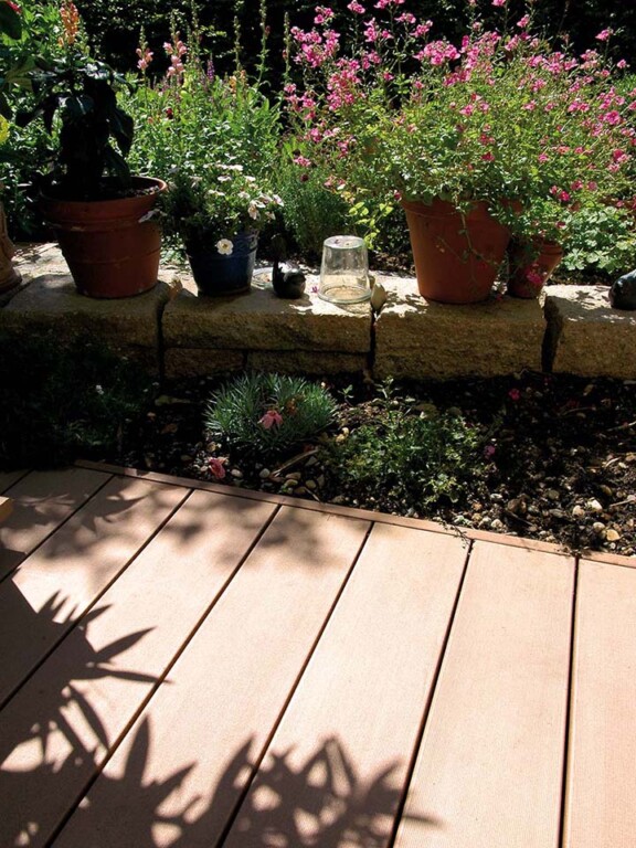 WPC Terrasse die an Blumenbeet und kleines Mäuerchen grenzt, auf dem bepflanzte Blumentöpfe stehen