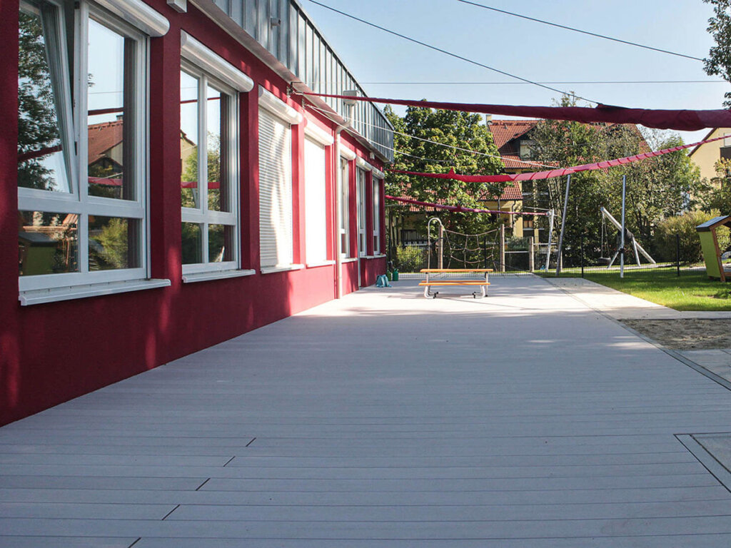 Vielfach ausgezeichnete graue Terrassendielen auf Außenfläche von Kita mit roter Fassade und Sonnensegeln über der Terrasse