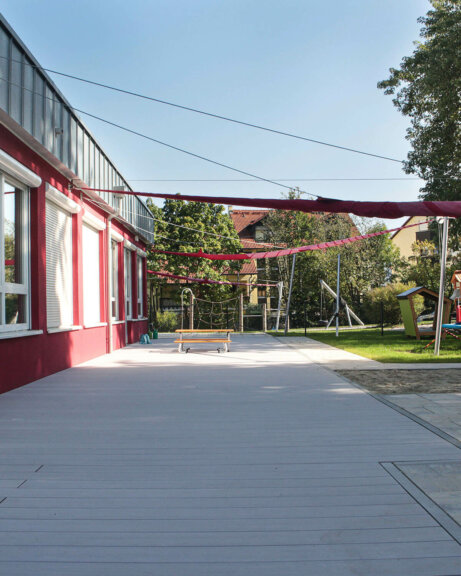 Vielfach ausgezeichnete graue Terrassendielen auf Außenfläche von Kita mit roter Fassade und Sonnensegeln über der Terrasse